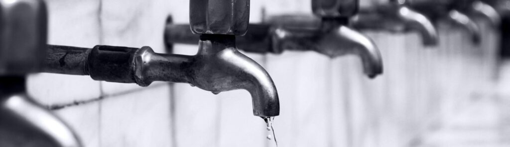 Contact Us Black and white image of a row of faucets with water dripping, highlighting plumbing details.